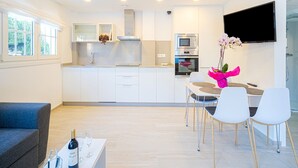 Una cocina moderna con armarios blancos, horno empotrado y microondas. Cuenta con un comedor con mesa y sillas, y un televisor montado en la pared.