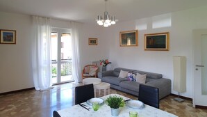 Una sala de estar con un sofá gris, una mesa de comedor con platos y una planta verde, una araña y una ventana con cortinas.