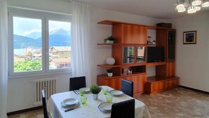 Un comedor con una mesa dispuesta para cuatro, una estantería de madera con un televisor, y vistas a un paisaje montañoso a través de amplios ventanales.