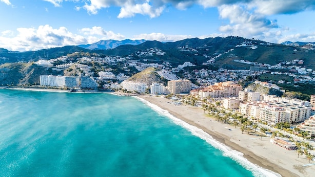 Un paisaje costero con playa, edificios y montañas al fondo.