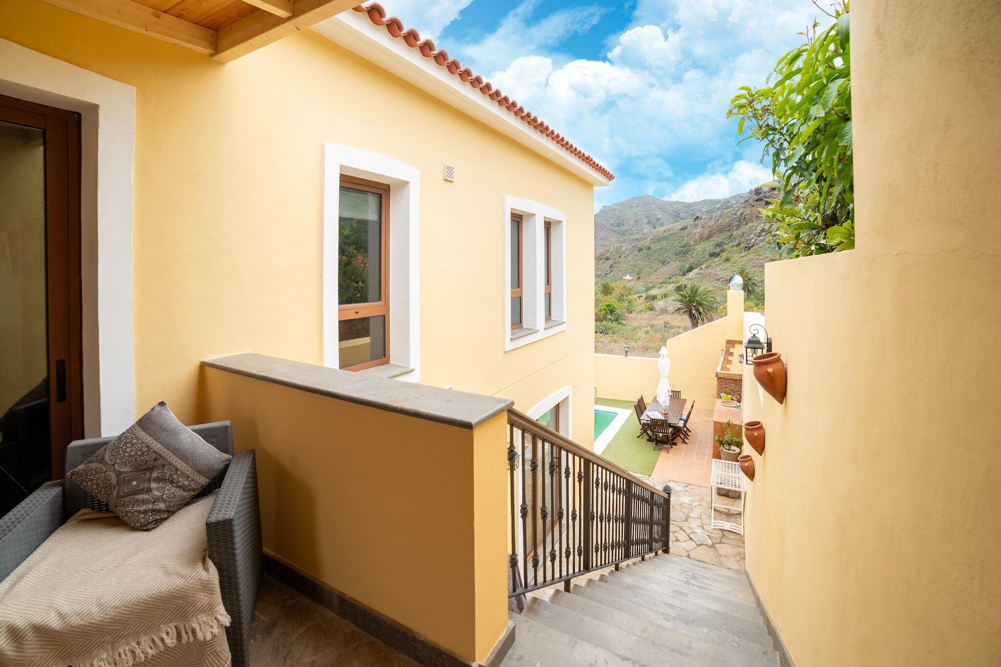 A balcony with a sofa and cushions, a small table with chairs, and a view of a mountainous landscape.