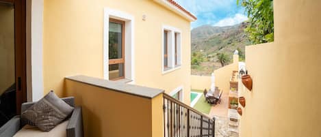 A balcony with a sofa and cushions, a small table with chairs, and a view of a mountainous landscape.