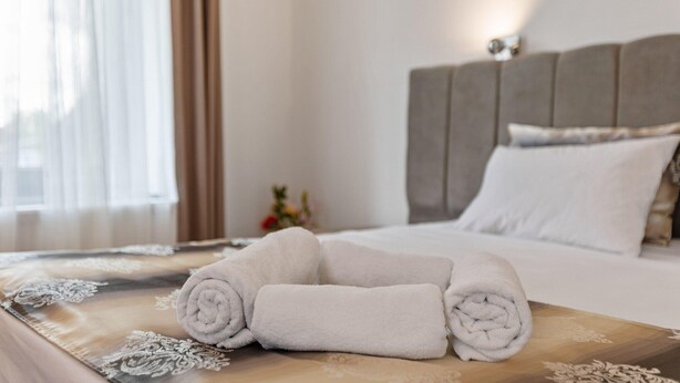 A neatly made bed with rolled white towels on a patterned bedspread, a headboard, and a bedside table with a vase of flowers.