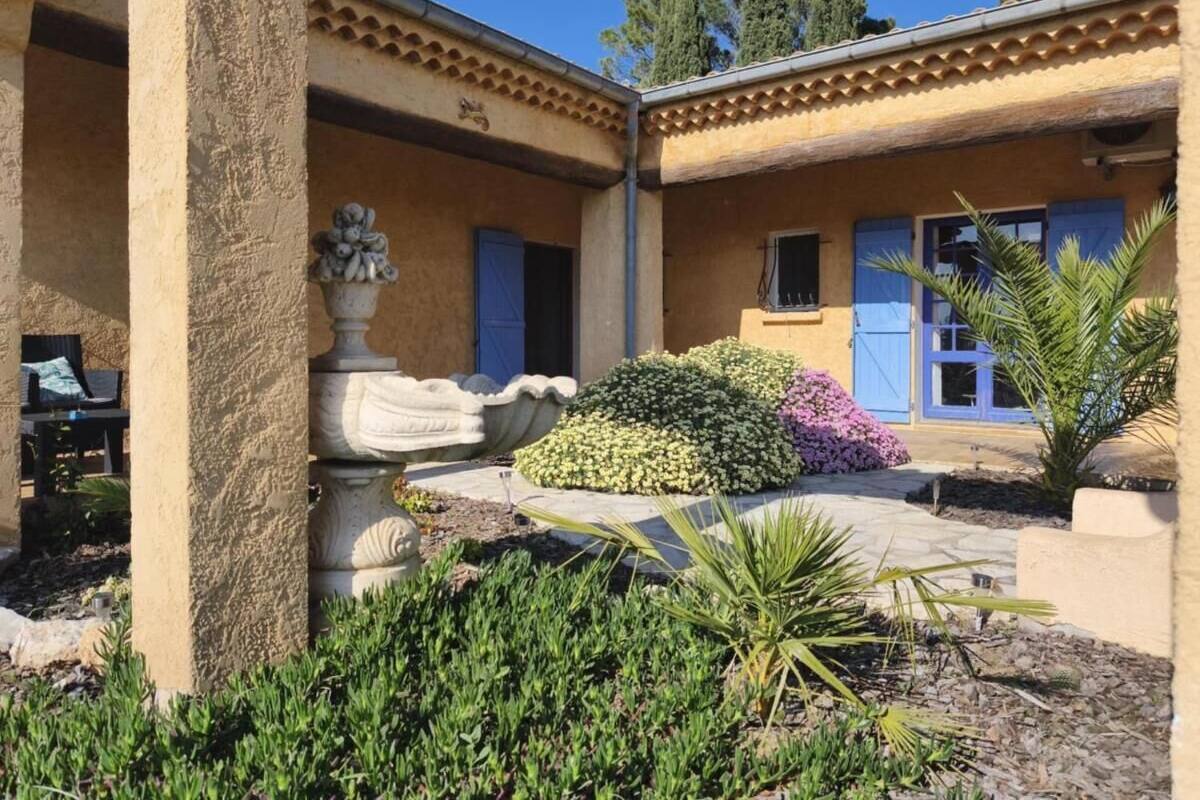 A courtyard with a stone fountain, a small garden with various plants, and a building with blue doors and a tiled roof.