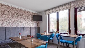 A dining area with wooden tables and blue chairs, a grey cushioned bench, and a TV mounted on the wall.