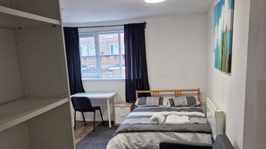 Quarto de hotel com cama, escrivaninha, cadeira, prateleira com ferro de passar roupa e um quadro na parede.