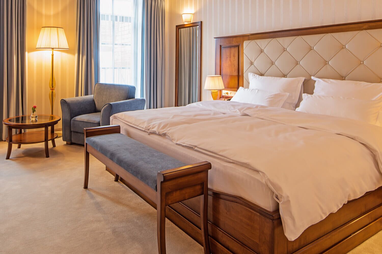 A hotel room with a large bed, a bench, a chair, two bedside tables, and a lamp.