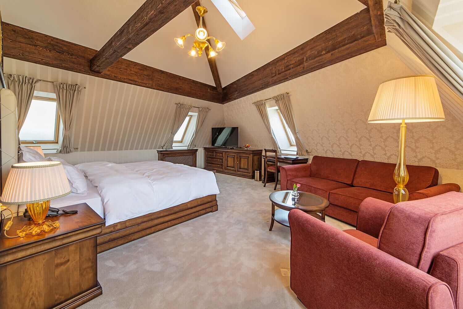 A hotel room with a large bed, a sofa, a desk, and a TV. There are two windows and a chandelier.