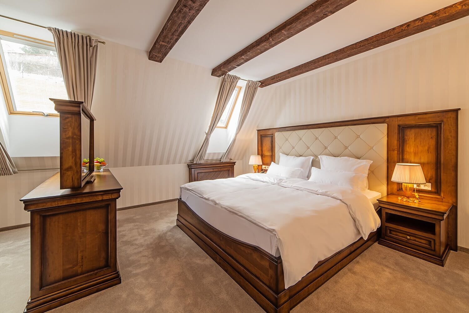 Un dormitorio con una cama grande, cabecera de madera, dos mesitas de noche con lámparas, cómoda de madera y una ventana con cortinas.