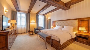 Ampia camera d'albergo con un letto grande, una testiera in legno, un angolo salotto con una poltrona, un tavolino e un televisore su un comò in legno.