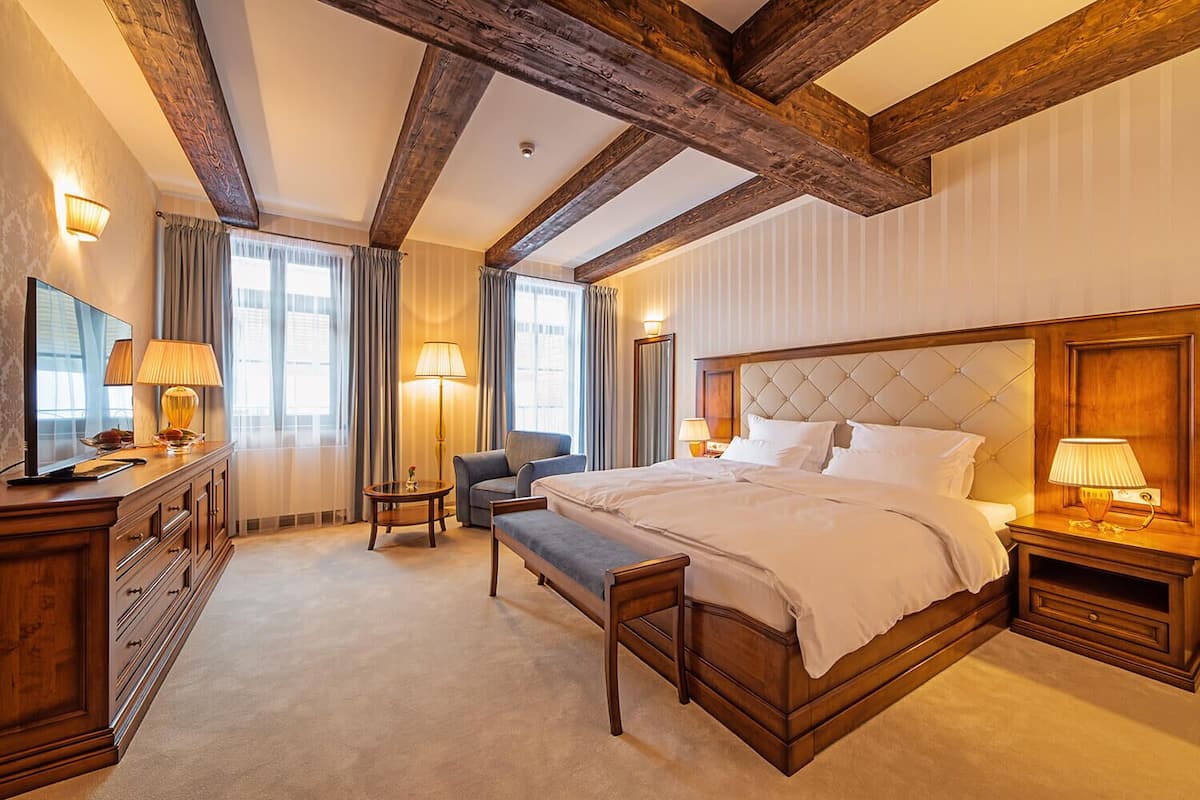 A spacious hotel room with a large bed, a wooden headboard, a sitting area with a chair, a small table, and a television on a wooden dresser.