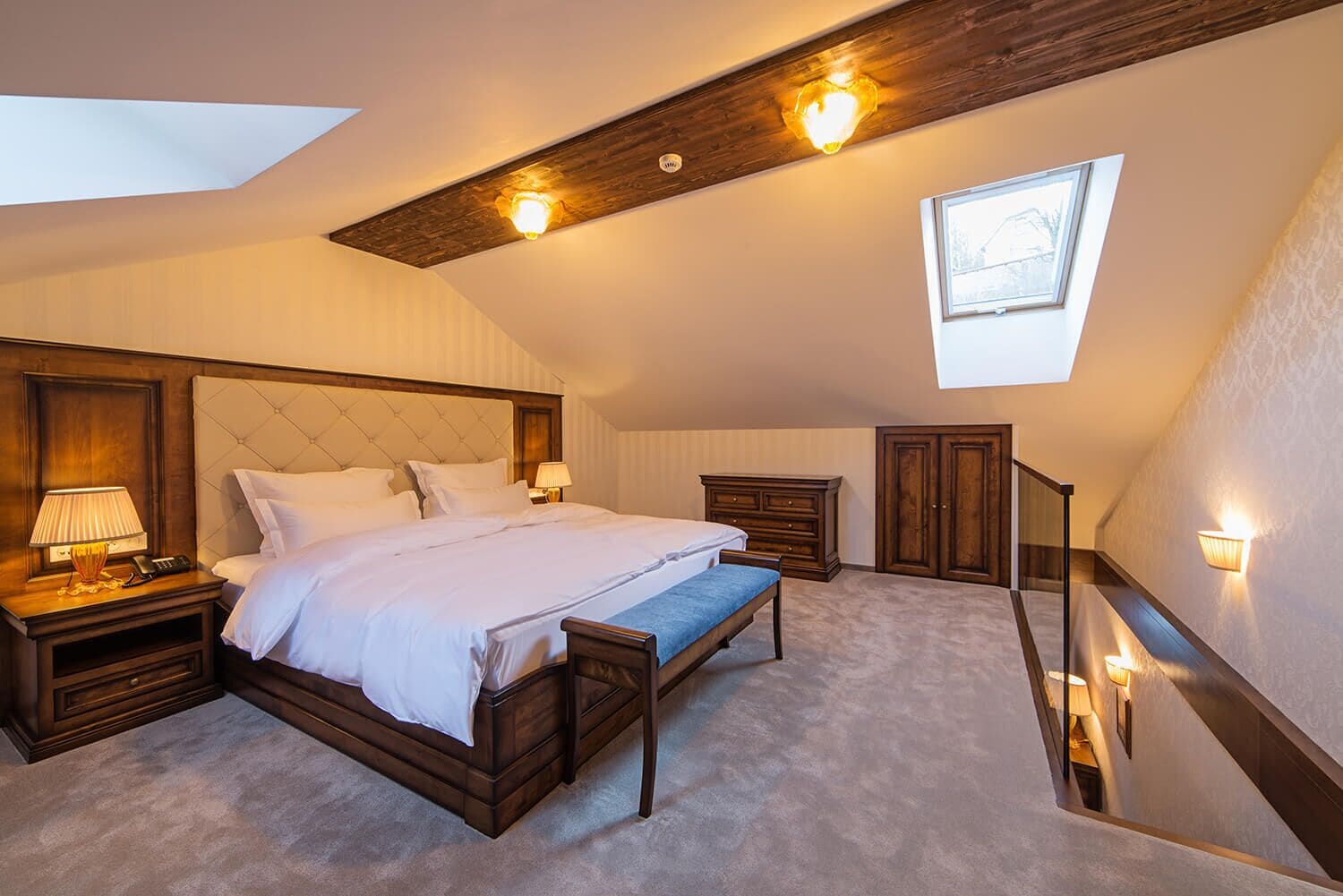Une chambre avec un grand lit, des tables de chevet, un banc, une armoire et une lucarne.