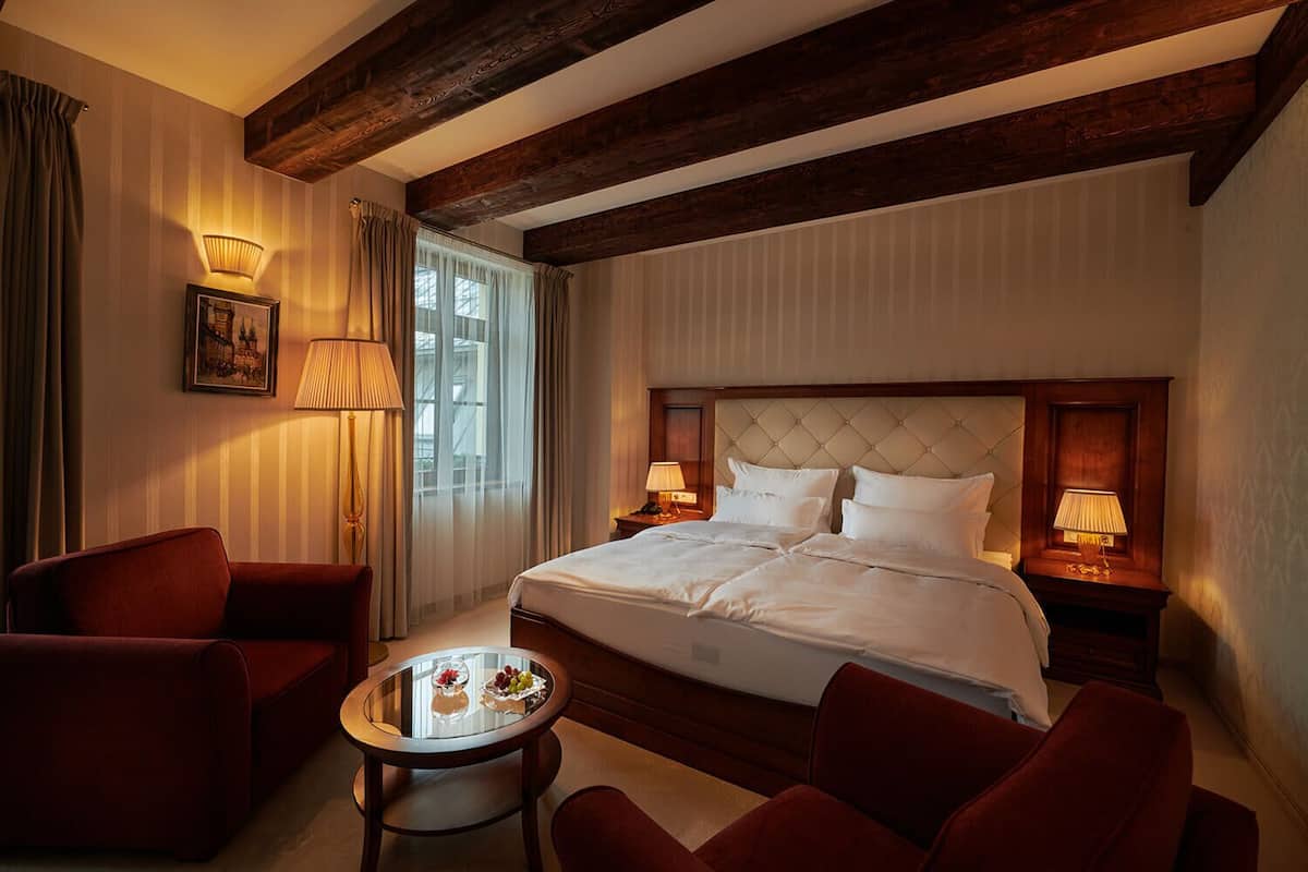 A hotel room with a large bed, two bedside tables with lamps, a small round table with a fruit bowl, and two armchairs.
