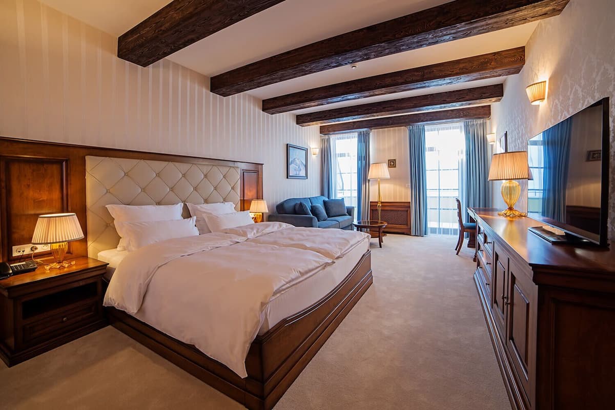 A hotel room with a large bed, wooden headboard, bedside lamps, a sofa, a TV, and a desk with a lamp.
