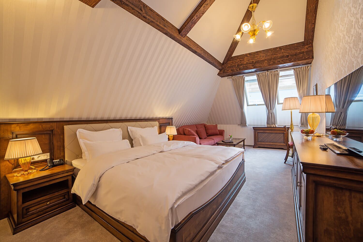 A spacious bedroom with a large bed, a wooden headboard, a nightstand with a lamp, a red sofa, and a desk with a lamp and fruit bowl.