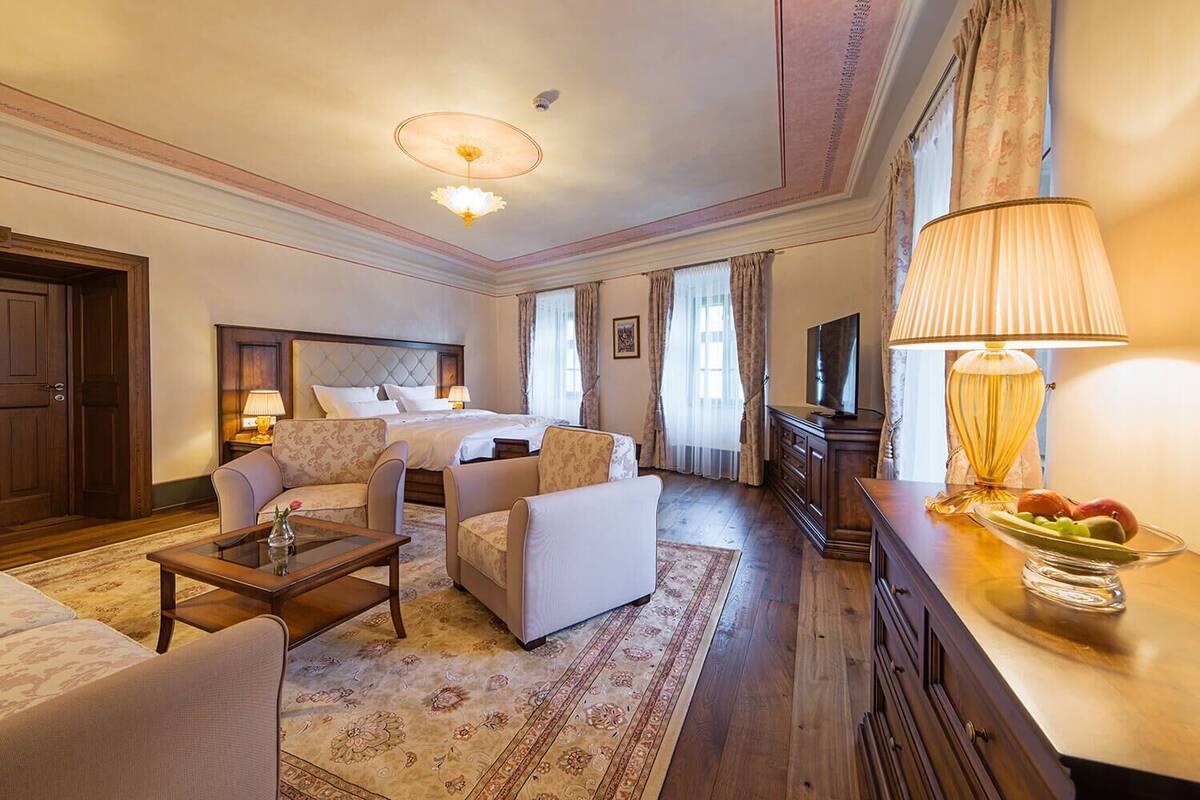 A hotel room with a bed, two armchairs, a sofa, a TV, a lamp, a fruit bowl, and a wooden dresser.
