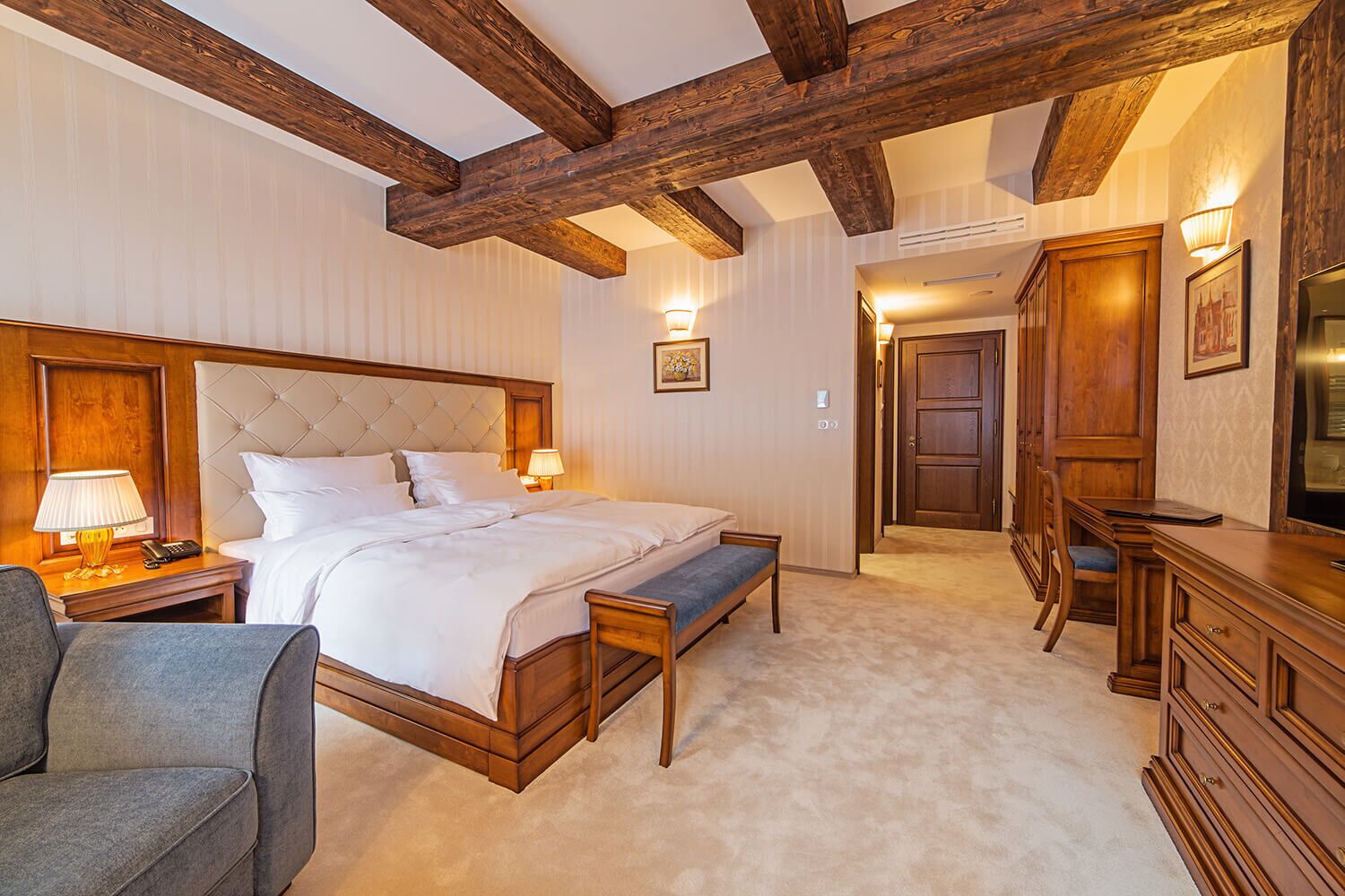 A hotel room with a large bed, bedside tables, a desk, a chair, and wooden beams on the ceiling.
