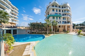 A modern hotel with a large swimming pool, multiple balconies, and palm trees.