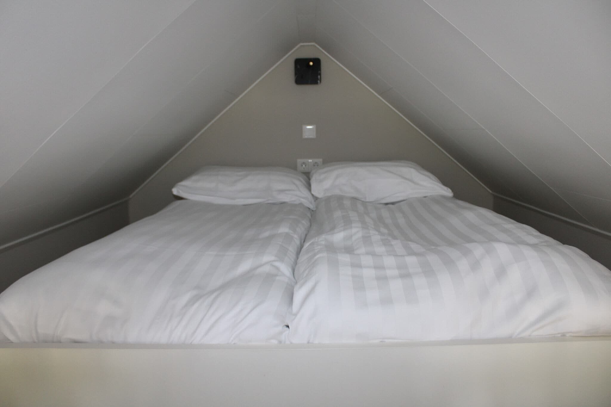 A small, triangular room with a single bed, a light switch, and a thermostat on the wall.