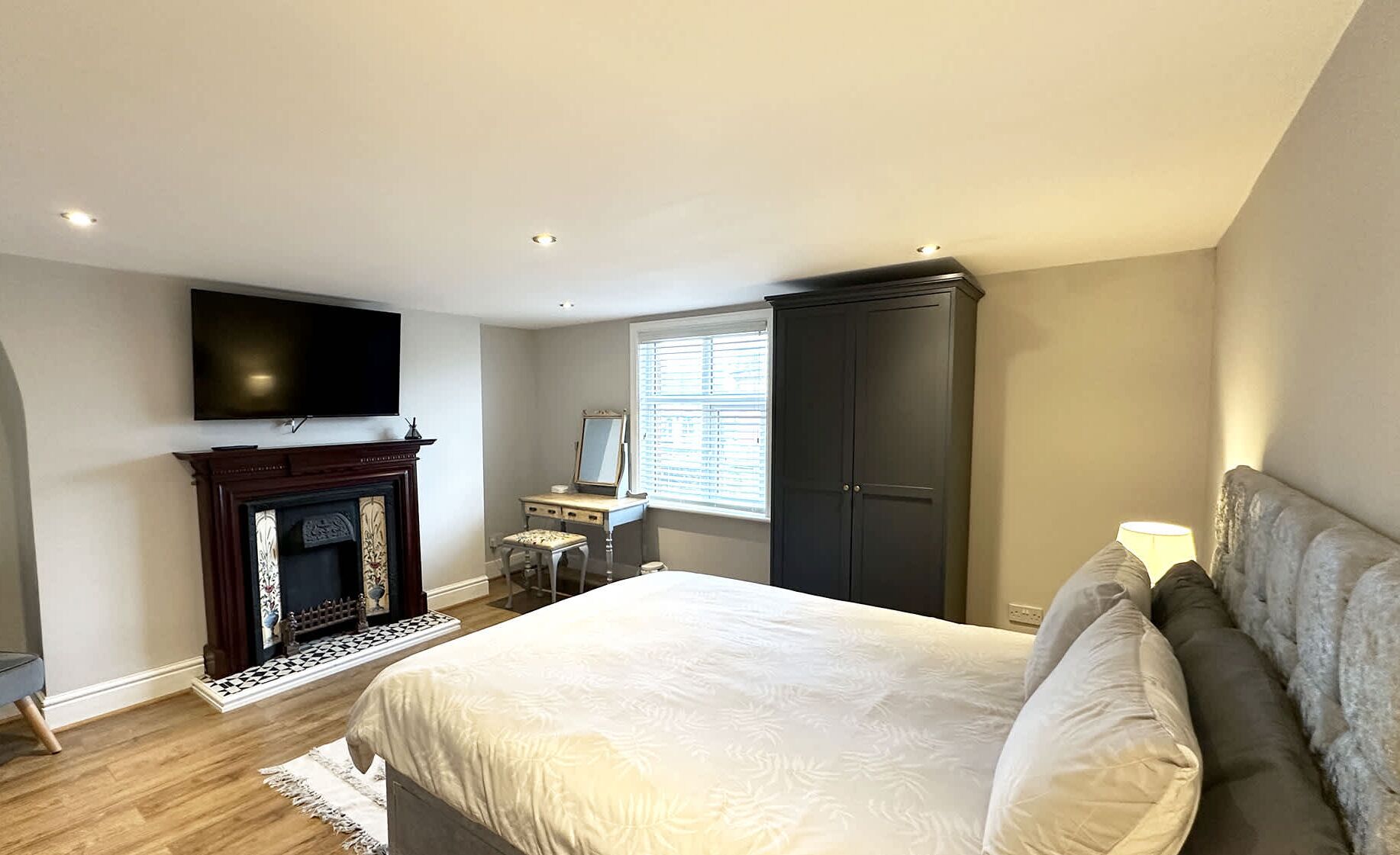 A bedroom with a bed, a TV mounted on the wall, a wardrobe, a desk with a chair, and a fireplace.
