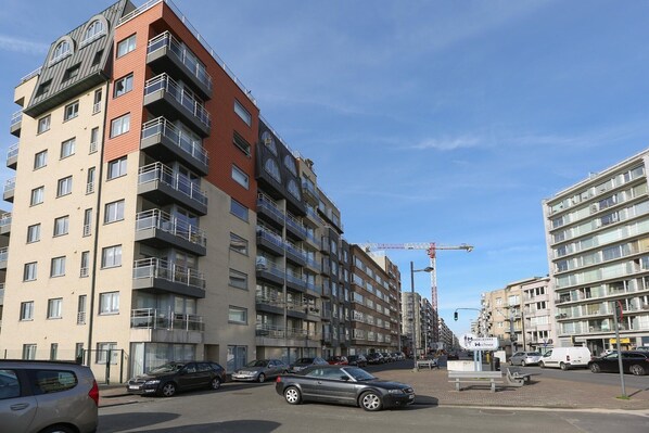 A multi-story residential building with balconies, parked cars, and a construction crane in the background.
