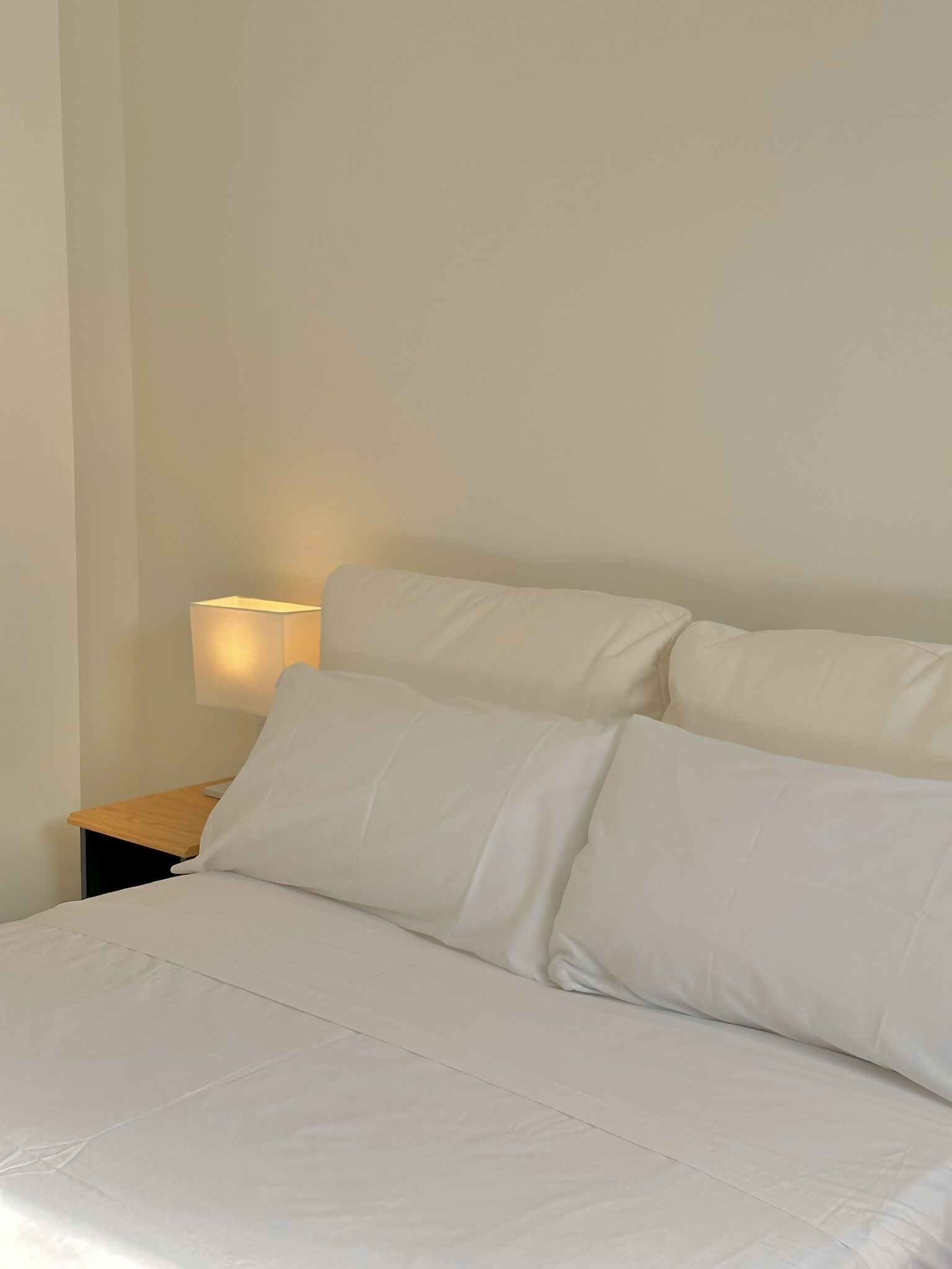 Un lit bien fait, avec du linge de lit blanc et des oreillers, une table de chevet avec une lampe et un mur sobre.