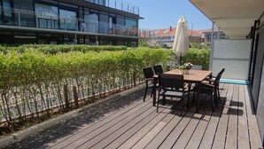 Een houten terras met een tafel en stoelen, omgeven door een haag en een modern gebouw op de achtergrond.