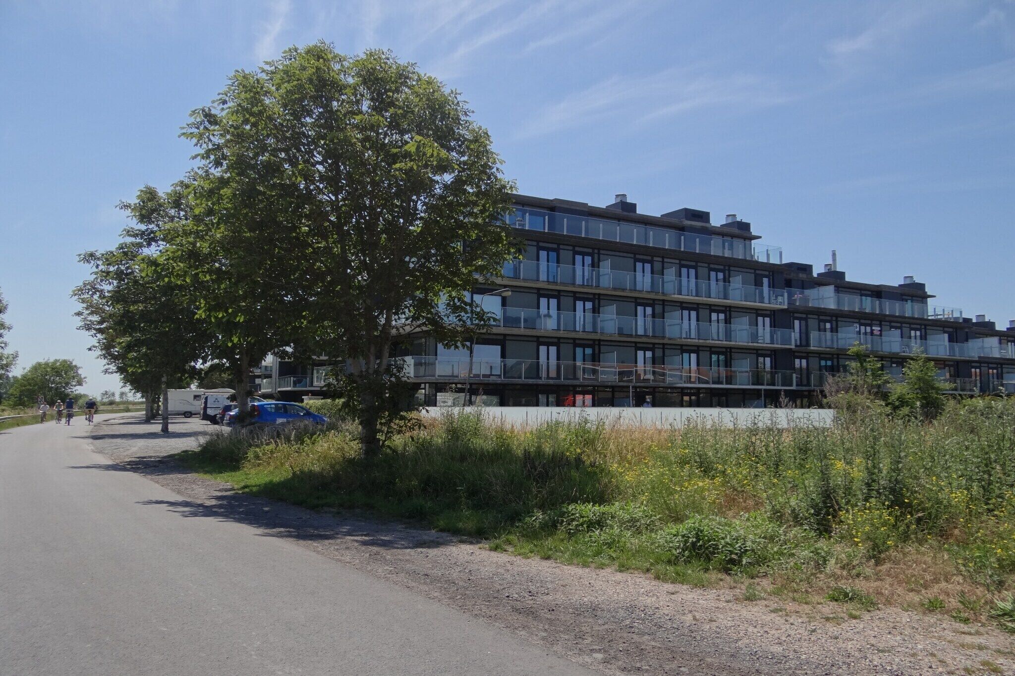 Een modern gebouw met meerdere verdiepingen en grote ramen, gelegen naast een weg met fietsers en een geparkeerde blauwe bestelwagen.