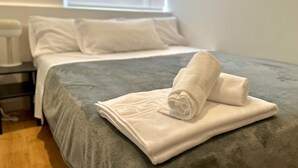 A neatly made bed with a grey duvet, white pillows, and a rolled white towel on top.