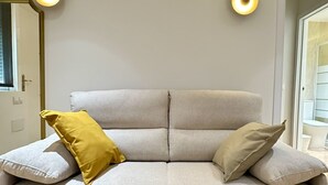 A light grey sofa with two cushions, one yellow and one beige, in a room with a wooden floor and two wall-mounted lights.