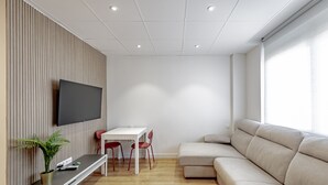 A modern living room with a flat-screen TV mounted on a wooden panel wall, a white table with red chairs, a beige sofa, and a potted plant.