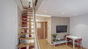 A modern apartment with a wooden staircase, a flat-screen TV, a dining area, and a small table with a plant.