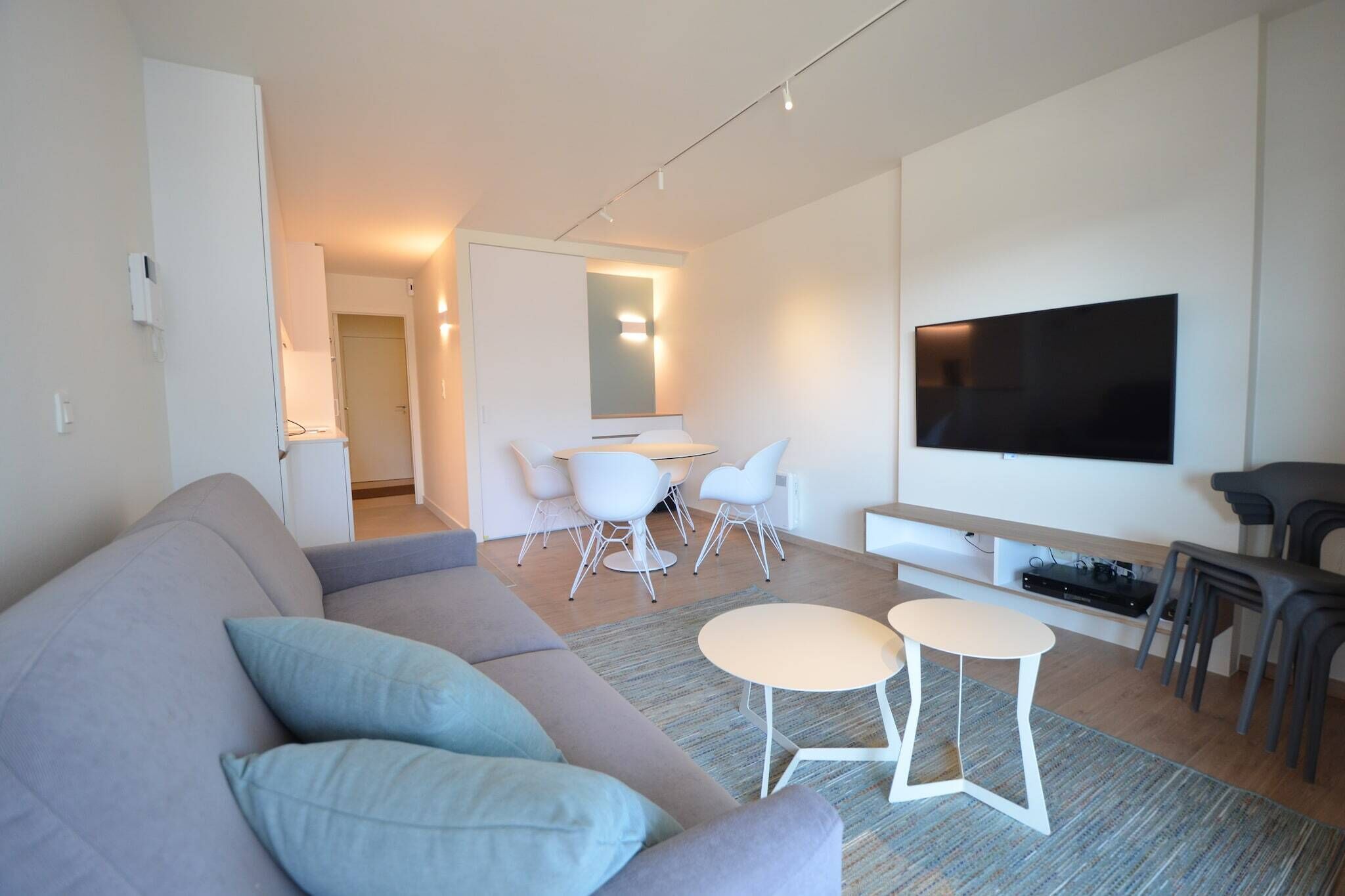 A modern living room with a grey sofa, a small round table, and a flat-screen TV mounted on the wall.
