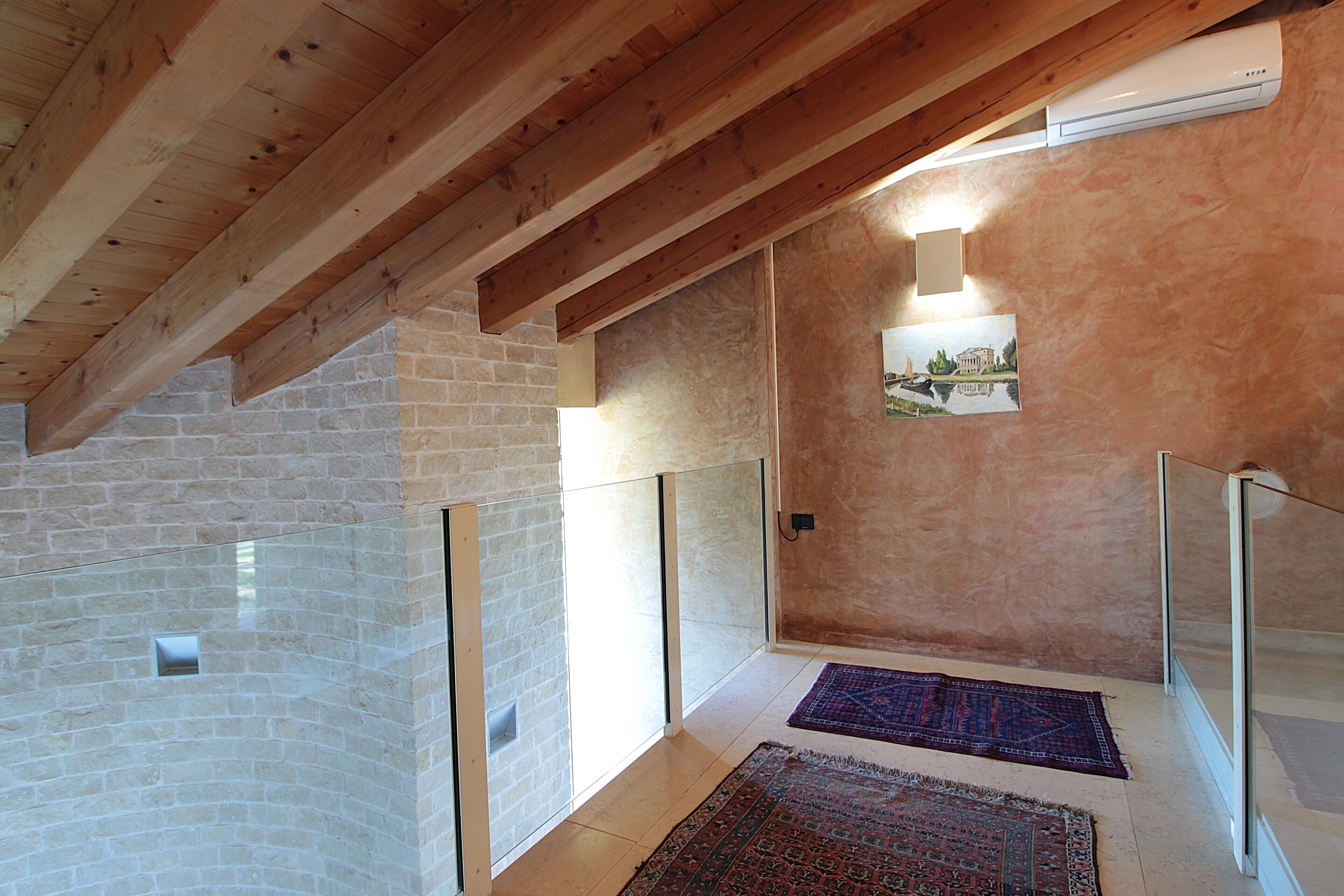 A high-ceilinged room with exposed wooden beams, a stone wall, and a glass railing.