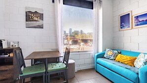 A room with a dining table, chairs, a sofa, a window with a city view, and a wall with framed pictures. - In the Heart of the City – Quaint Studio in the Prime Location! (Melbourne)