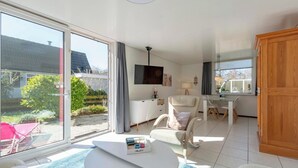 En rummelig opholdsstue med en stor skydedør, der fører til en have, en hvid sofa, et lille hvidt bord og et træskab.