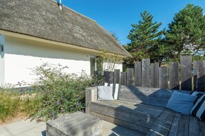 Una terraza de madera con una casa de techo de paja, una valla de madera y un banco con cojines.