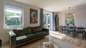 A modern living room with a sofa, dining table, and chairs. There is a painting on the wall and a window with curtains.