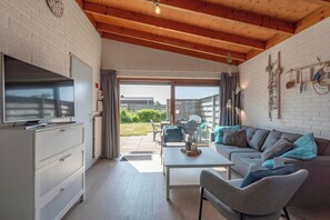 A modern living room with a TV, sofa, and dining area, featuring a wooden ceiling and large windows.