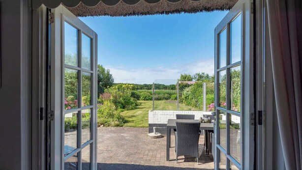 Eine offene Tür führt zu einer Terrasse mit Tisch und Stühlen, einem grünen Garten und blauem Himmel.