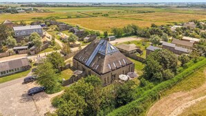 An aerial view of a rural property with multiple buildings, including a prominent house with a sloped roof and skylights, surrounded by open fields and sparse vegetation. - Vossehoeck 3 Apartment with nature reserve view (Callantsoog)