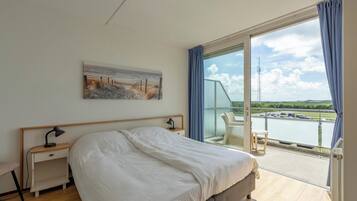 Een hotelkamer met een groot bed, een nachtkastje met lamp, een stoel en uitzicht op een waterpartij vanaf een balkon.
