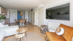 A modern living room with a sofa, ottoman, and coffee table, a dining area with chairs, and a kitchen in the background. - Apartment in Groote Keeten near the Beach (Groote Keeten)
