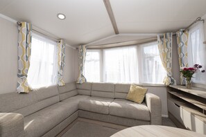 Cozy living area with a sectional sofa, round table, and wooden cabinet, featuring a curved ceiling and two windows with patterned curtains.