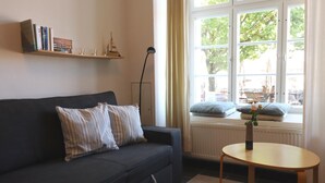 A living room with a dark sofa, striped pillows, a small round table with a vase, and a window with a view of trees and buildings.