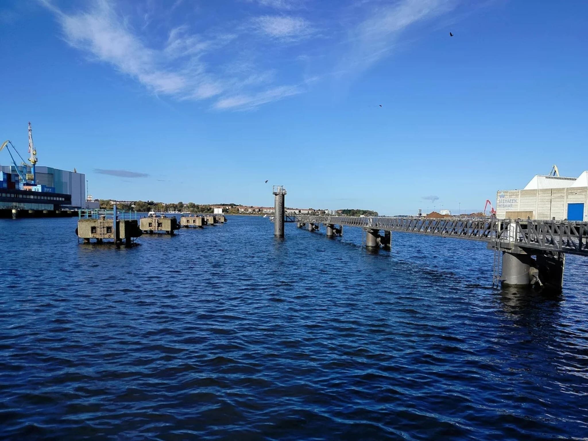 Ein Ufer mit einer Pier, die sich ins Wasser erstreckt, ein großes Gebäude mit einer blauen Tür und ein Kran im Hintergrund.