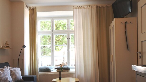A compact living space with a sofa, a small table, a radiator, a window with curtains, and a television mounted on the wall.