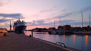 A waterfront marina at dusk with a large ship docked on the left and several smaller boats in the background.