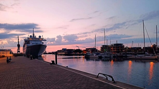 A waterfront marina at dusk with a large ship docked on the left and several smaller boats in the background.