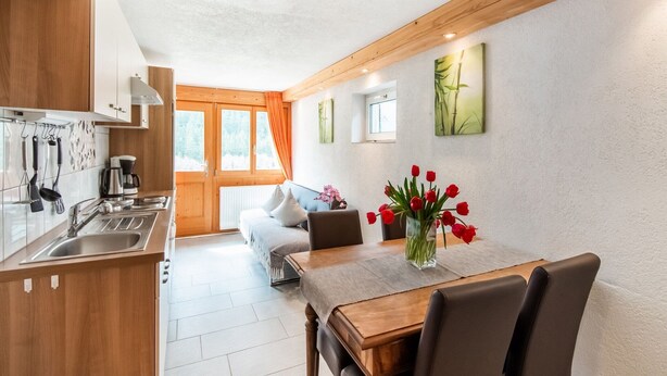 A modern kitchen with wooden cabinets, a dining area with a table and chairs, and a view of a snowy landscape through large windows. - Apartment in Sankt Gallenkirch near Ski Slopes (Vorarlberg)